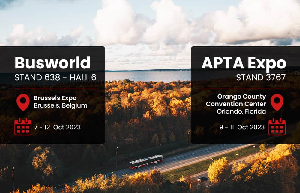 Red bus driving down a highway with forest either side while two signs read Busworld and APTA Expo